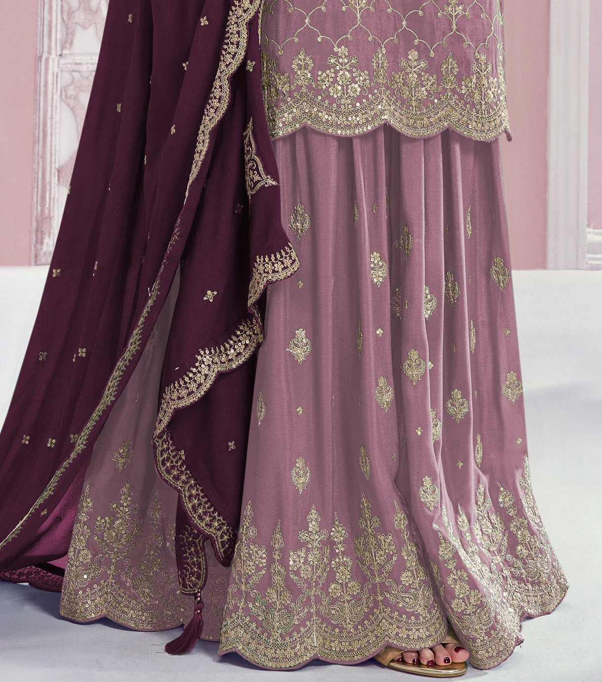 A close-up of a traditional outfit featuring a dusty pink embroidered skirt with gold detailing, paired with a deep purple dupatta and matching embroidered border, shown on a model.
