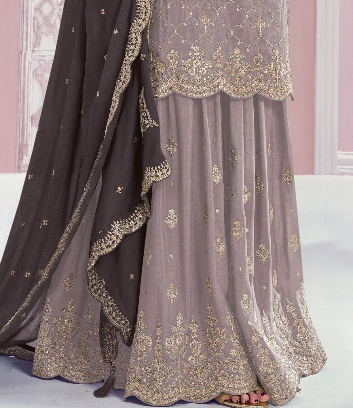 A close-up of a traditional Indian outfit featuring a dusty purple skirt with gold embroidery, a matching embroidered dupatta, and a dark brown border, shown on a model