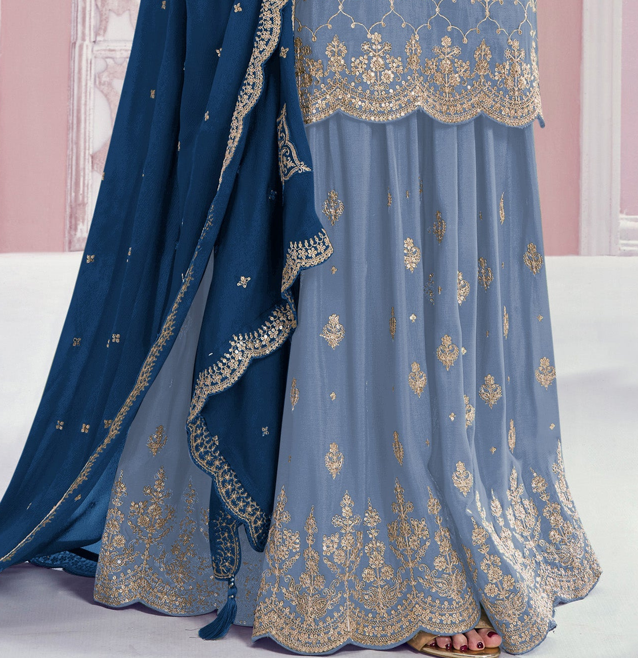 A close-up of a traditional Indian outfit featuring a light blue skirt with intricate gold embroidery, a matching blue dupatta, and a blue top with gold border detailing. A person's feet are visible at the bottom.